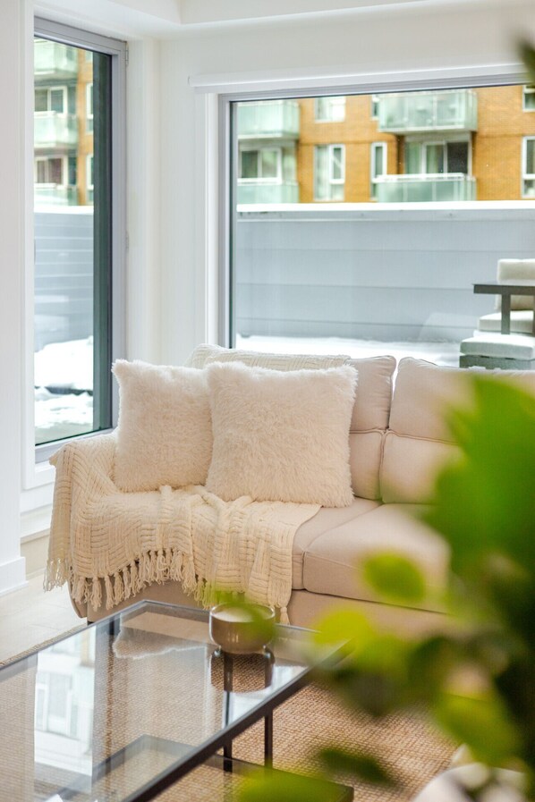 A modern living room with a beige sofa, a glass coffee table, and a view of a building through large windows. - Premium Living | Rooftop +Underground Parking (Montreal)