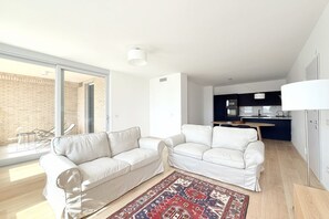 Ein modernes Wohnzimmer mit zwei weißen Sofas, einem gemusterten Teppich und einer Küche im Hintergrund.