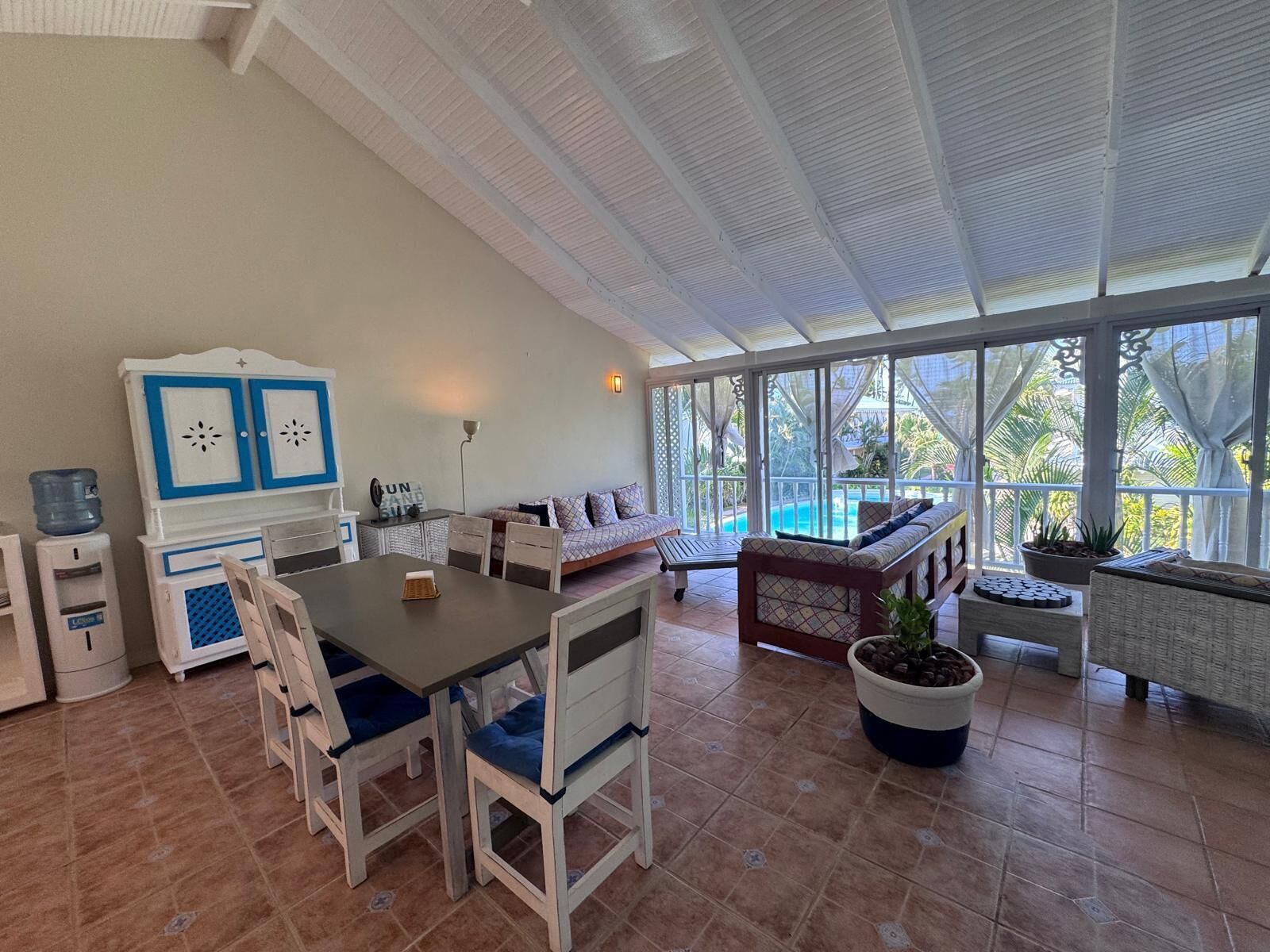 A spacious room with a large dining table, chairs, a sofa, and a view of a swimming pool through large windows.