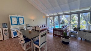 A spacious room with a large dining table, chairs, a sofa, and a view of a swimming pool through large windows. - Spacious 3-bedroom condo in adorable Las Terrenas with AC (Las Terrenas)