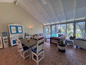 A spacious room with a large dining table, chairs, a sofa, and a view of a swimming pool through large windows. - Spacious 3-bedroom condo in adorable Las Terrenas with AC (Las Terrenas)