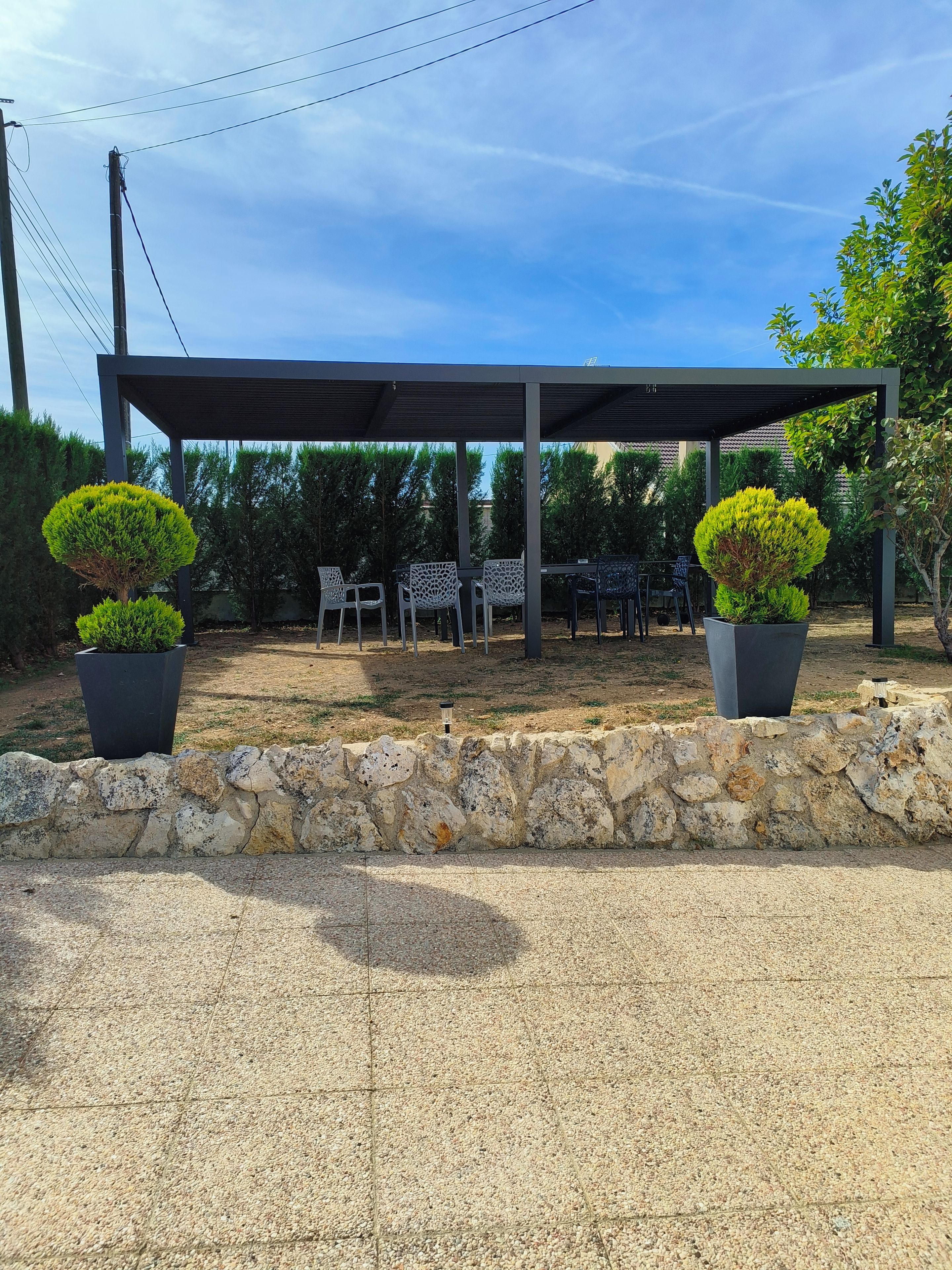 A covered outdoor seating area with tables and chairs, surrounded by a stone wall and potted plants.