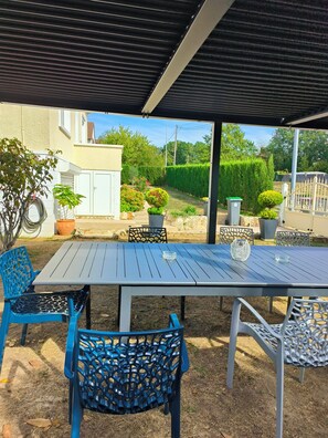 An outdoor patio with a table and chairs, surrounded by a garden and a white building.