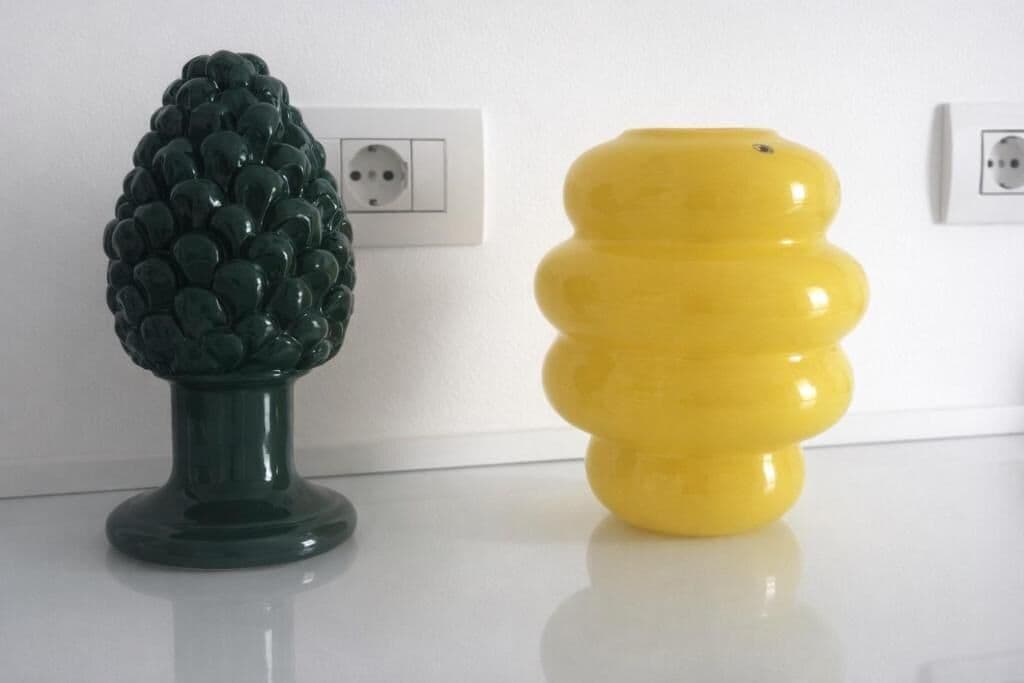 Deux vases décoratifs sont posés sur une surface blanche, devant un mur blanc et sur lequel on aperçoit des prises électriques à l’arrière-plan.