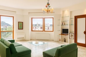 En stue med en grøn sofa, et glasbord, et fjernsyn på en hylde og udsigt til bygninger gennem store vinduer.