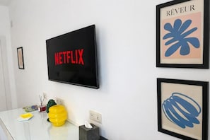 A wall-mounted television with the word 'NETFLIX' in red letters, a framed artwork with the word 'RÊVEUR' and a blue abstract design, and a small table with a yellow object and a tissue box. - roof and sky (Sorrento)