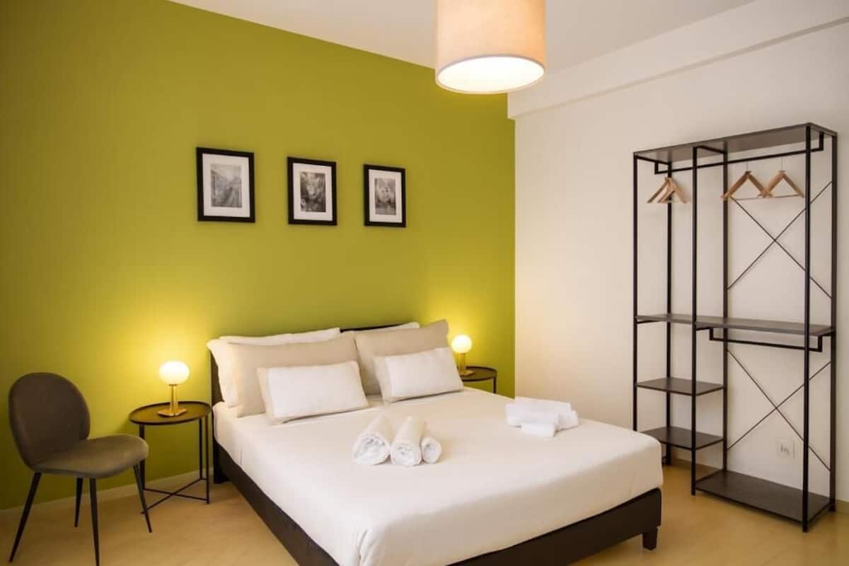 A hotel room with a bed, a chair, a bedside table, a lamp, a wardrobe, and three framed pictures on the wall.
