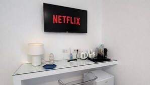 Una habitación de hotel moderna con un escritorio de vidrio, un refrigerador pequeño, un cesto de basura, una cafetera, un hervidor de agua, una lámpara y un televisor que muestra el logotipo de Netflix.