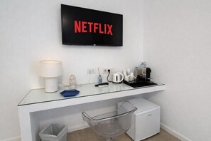 Una habitación de hotel moderna con un escritorio de vidrio, un refrigerador pequeño, un cesto de basura, una cafetera, un hervidor de agua, una lámpara y un televisor que muestra el logotipo de Netflix.