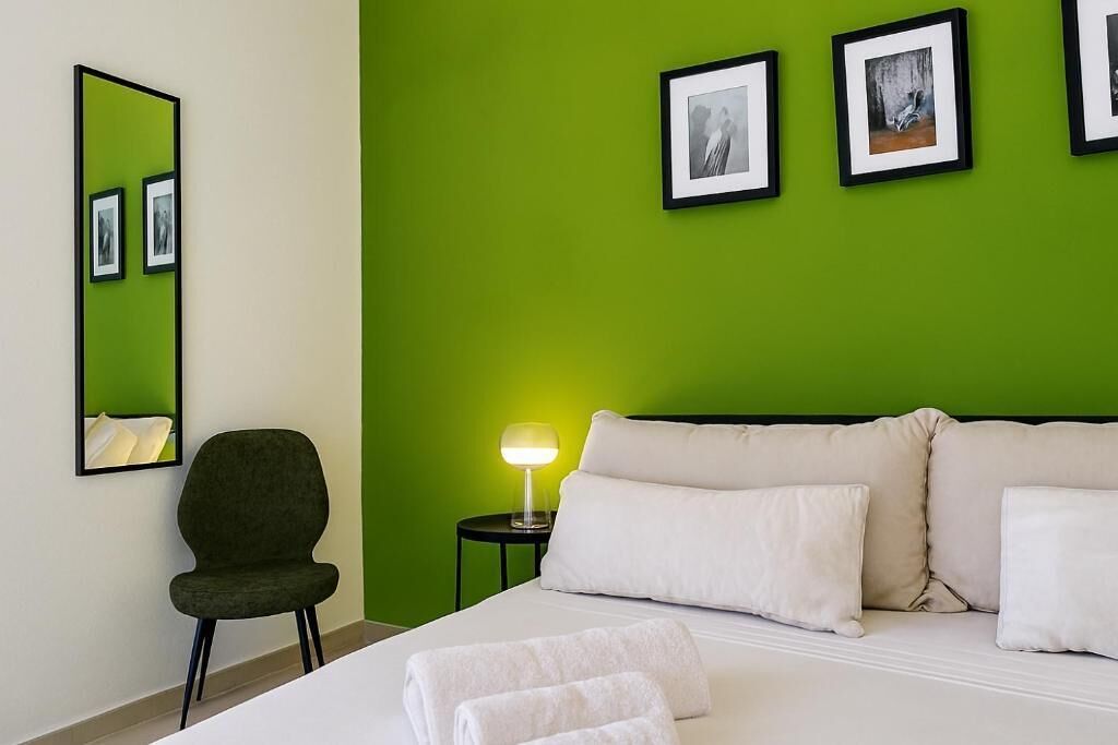 A hotel room with a green wall, a bed with white linens, a chair, a bedside table with a lamp, and framed pictures on the wall.