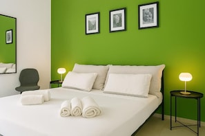 Hotellihuone, jossa on vihreä seinä, valkoisilla lakanoiden peitetty sänky, kaksi yöpöytää valaisimineen ja kolme kehystettyä kuvaa seinällä.