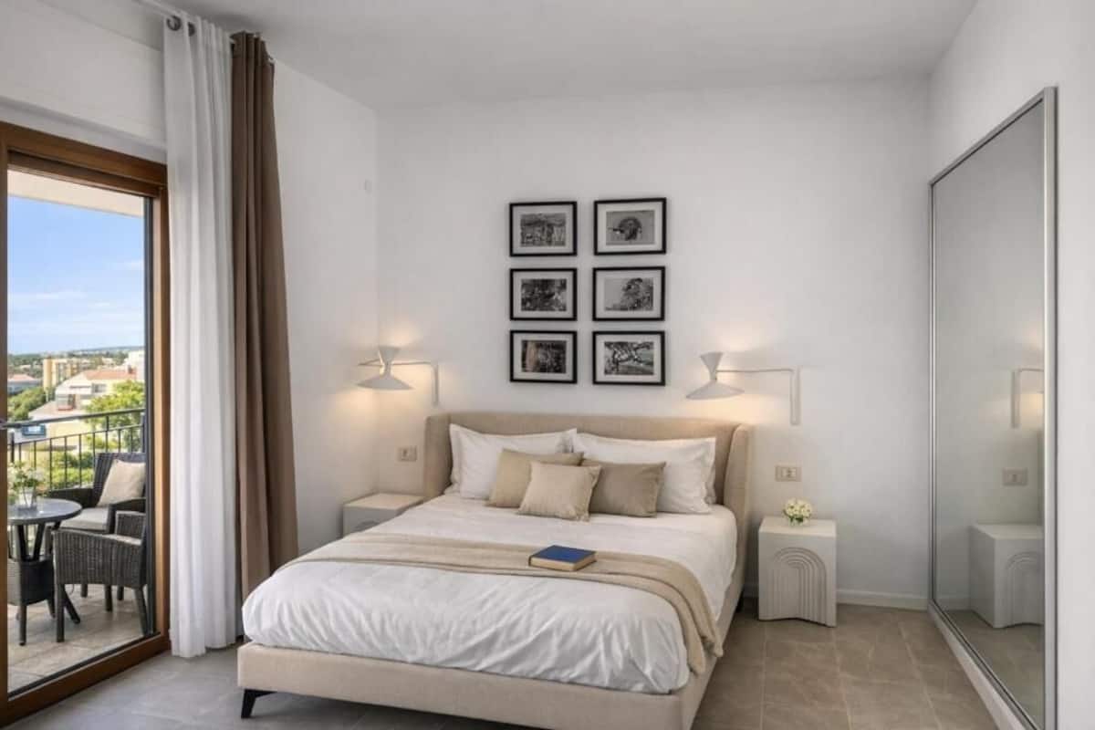 A bedroom with a bed, a balcony with outdoor furniture, and framed pictures on the wall.