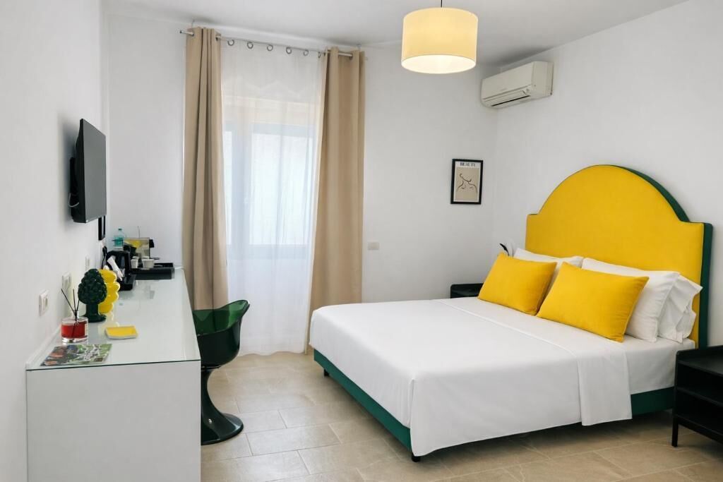 A hotel room with a bed, a desk, a chair, a TV, a window with curtains, and an air conditioning unit.