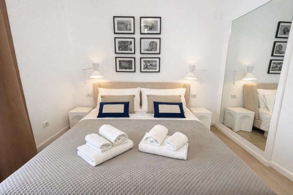 Un lit bien fait, agrémenté de serviettes, d’une tête de lit, de tables de chevet, de lampes fixées au mur et de tableaux encadrés.