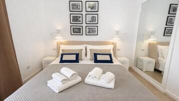 Un lit bien fait, agrémenté de serviettes, d’une tête de lit, de tables de chevet, de lampes fixées au mur et de tableaux encadrés.