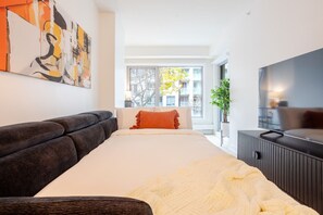 Un dormitorio moderno con una cama, un sofá oscuro, un televisor y una ventana con vistas a edificios y árboles.