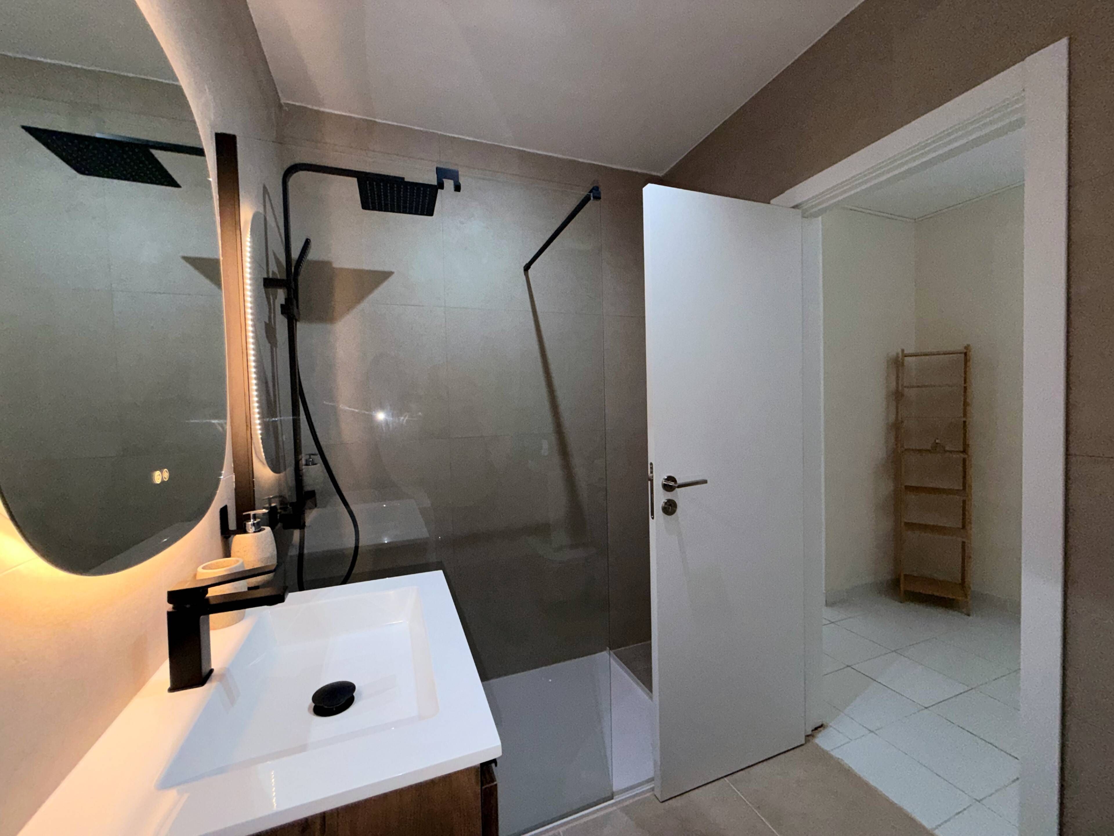 Un baño con lavamanos blanco, un espejo, una ducha y una puerta que da a otra habitación.