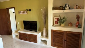 Un salon avec un téléviseur posé sur un meuble, une armoire en bois, une étagère ornée d’objets décoratifs et une porte donnant sur une autre pièce.