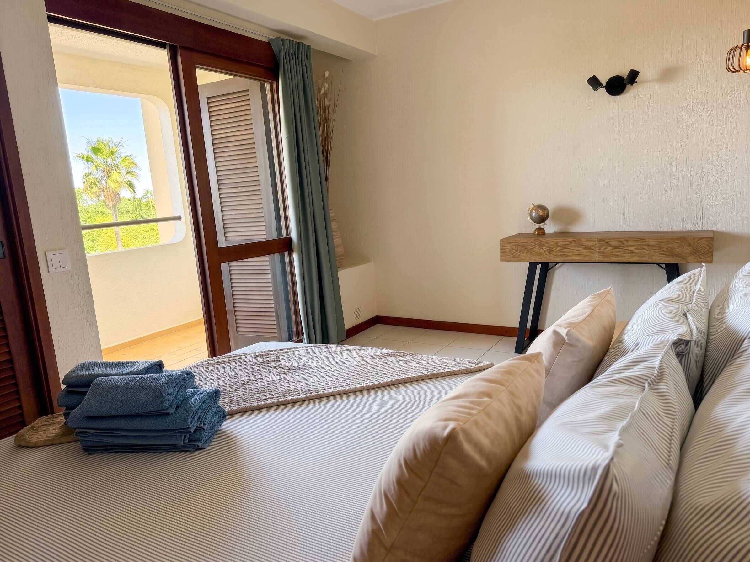 A bedroom with a bed, pillows, a bedside table with a globe, a view through a window with blinds, and a door leading to an outdoor balcony.