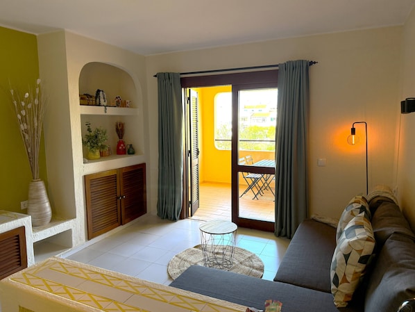 A living room with a sofa, a small round table, a balcony with a table and chairs, and a wall-mounted lamp. - Authentic apartment, 2 lovely private balconies (Albufeira)