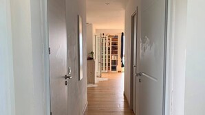 Een gang met houten vloer en witte deuren, die leidt naar een kamer met een boekenkast en een raam.