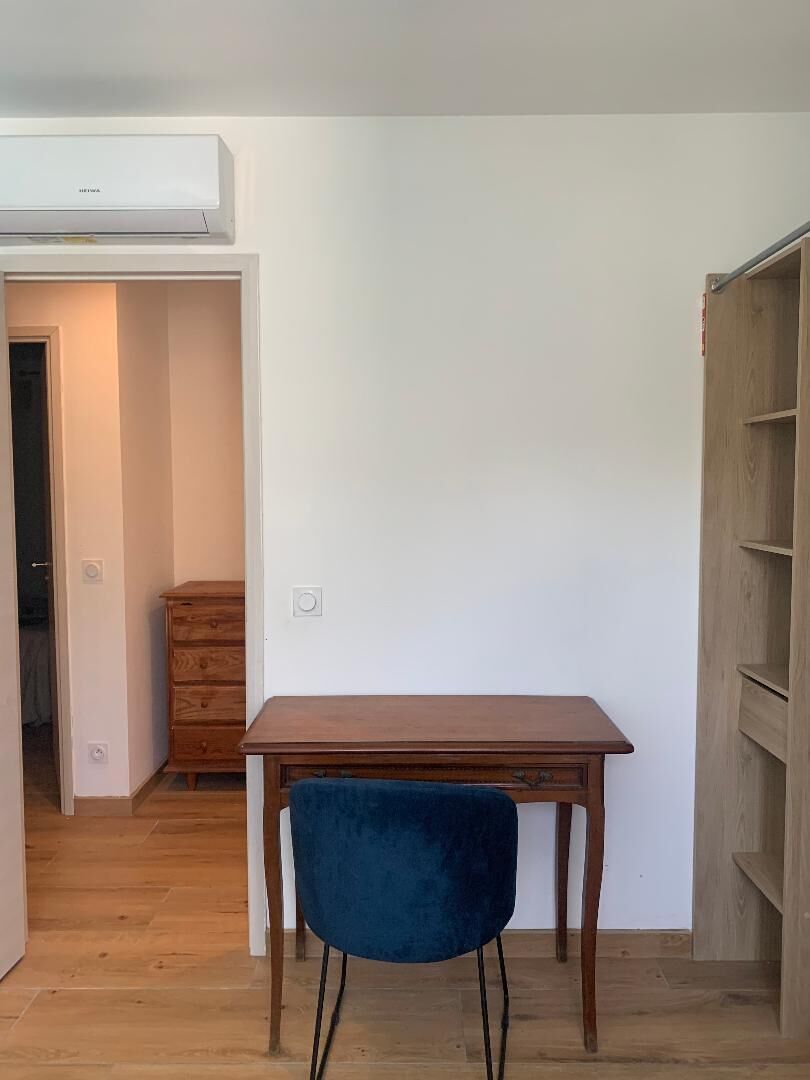 A room with a wooden desk, a blue chair, a wardrobe, and an air conditioning unit.