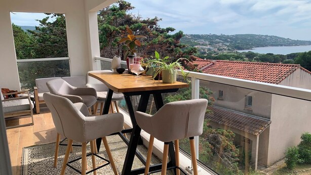 Een balkon met een tafel, stoelen, een bloempot en uitzicht op een kustlandschap.