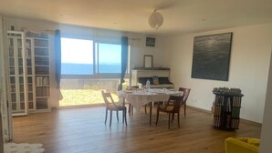Een eetgedeelte met een tafel voor vier personen, een boekenkast, een piano en uitzicht op zee.