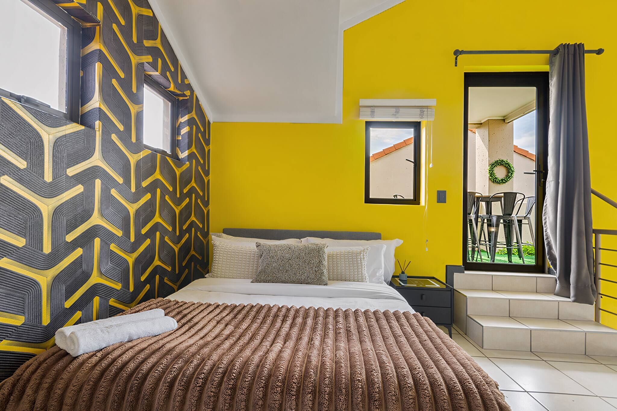 Camera da letto con parete decorata, un letto con coperta marrone testurizzata, un comodino e vista su un balcone con sedie.