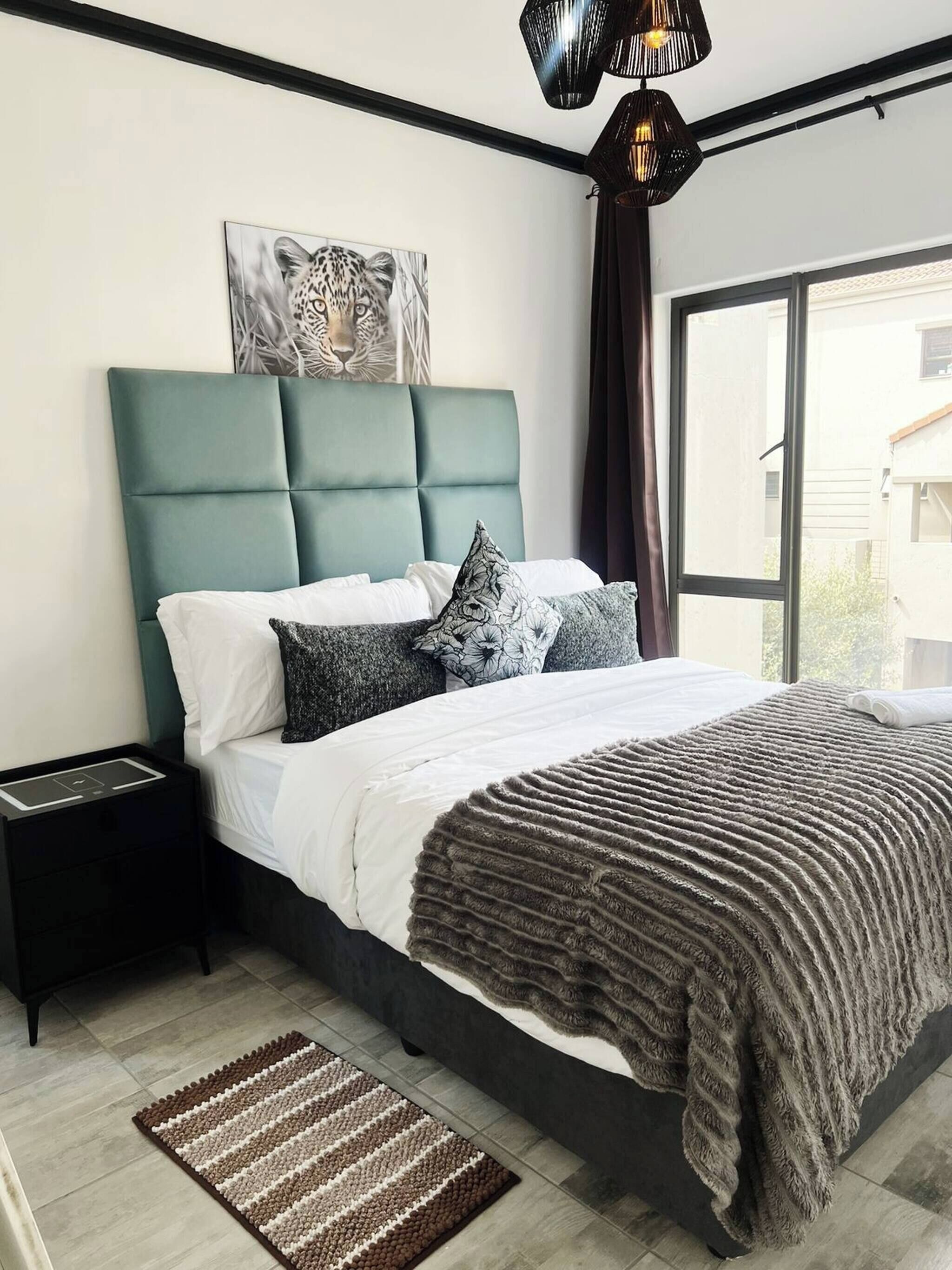 Un dormitorio moderno con un cabecero verde azulado, un ventanal grande, una mesita de noche negra y una alfombra estampada.