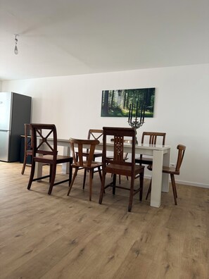 Un coin repas avec une table blanche et des chaises en bois, un réfrigérateur noir et une décoration murale sur le thème de la forêt.