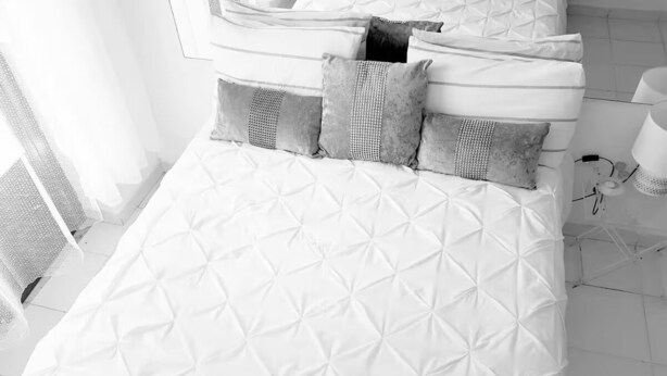 A neatly made bed with a quilted pattern, a bedside table with a lamp, and a window with curtains. - Delgado Airport Suite (Lisbon)