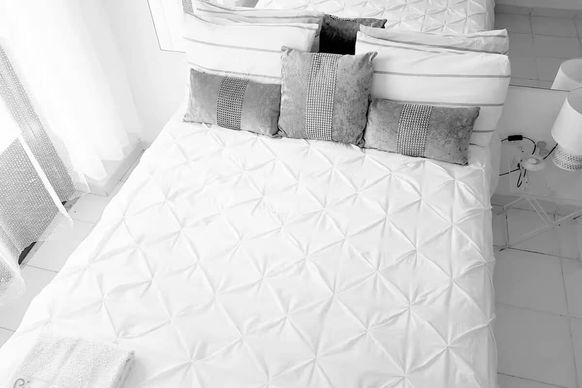 A neatly made bed with a quilted pattern, a bedside table with a lamp, and a window with curtains.