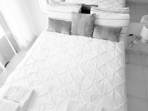 A neatly made bed with a quilted pattern, a bedside table with a lamp, and a window with curtains. - Delgado Airport Suite (Lisbon)