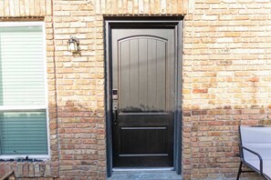 Ein Backsteinhaus mit einer dunklen Holztür, einem Fenster mit Rollläden und einem Metallstuhl davor.