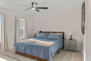 Ein Schlafzimmer mit Bett, Nachttischen, einem Deckenventilator und einem Fenster mit Vorhängen.