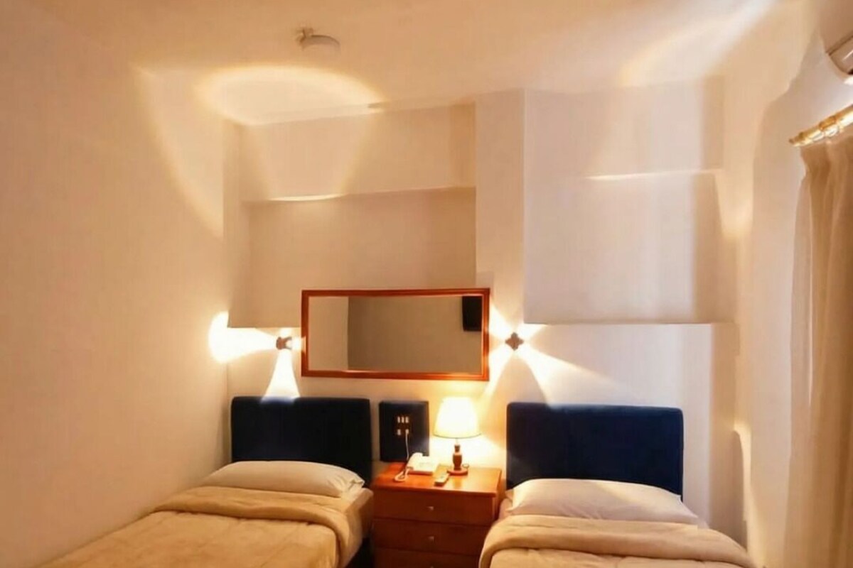 A hotel room with two single beds, a bedside table with a lamp, a mirror, and a wall-mounted light fixture.