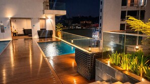 Ein Poolbereich auf dem Dach mit Holzterrasse, Glasgeländer und einer Sitzgruppe mit Stühlen.
