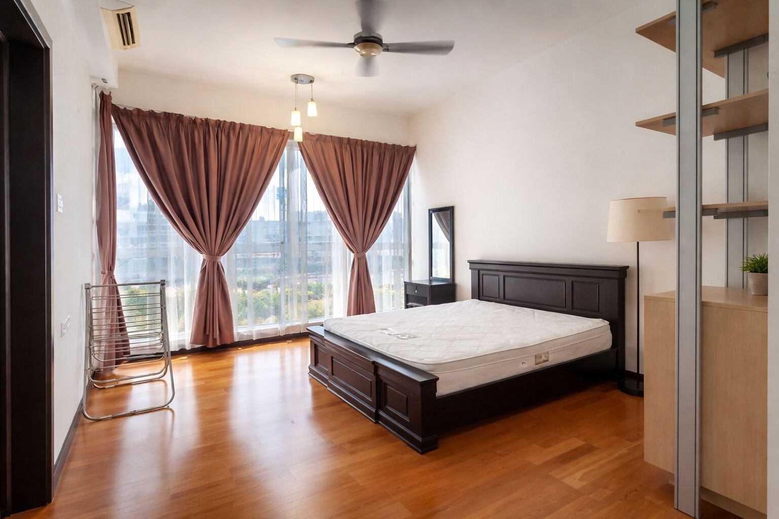 A bedroom with a bed, a ceiling fan, a window with curtains, a nightstand, and a view of the city.