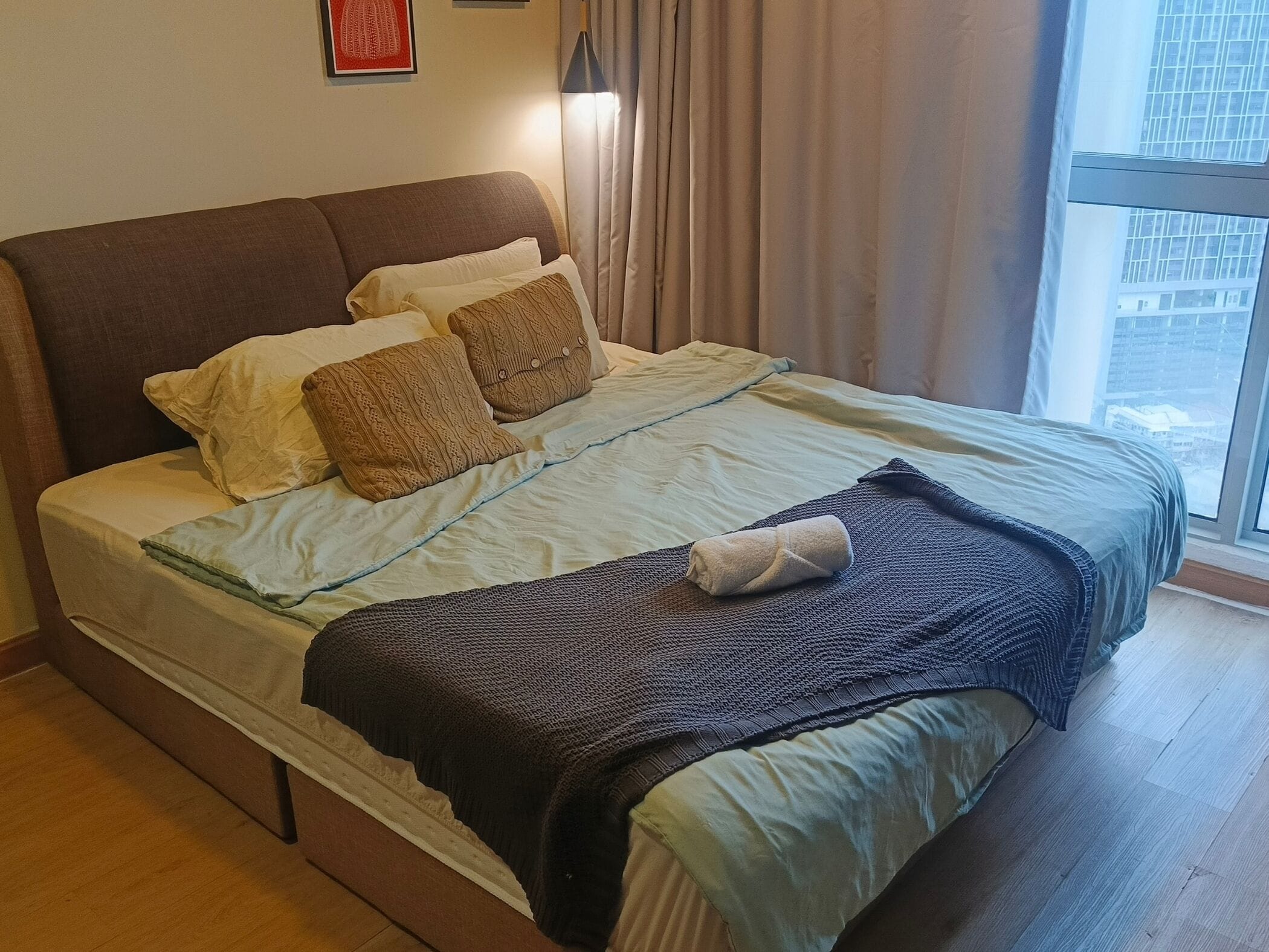 A bed with a headboard, pillows, a blanket, and a rolled towel.