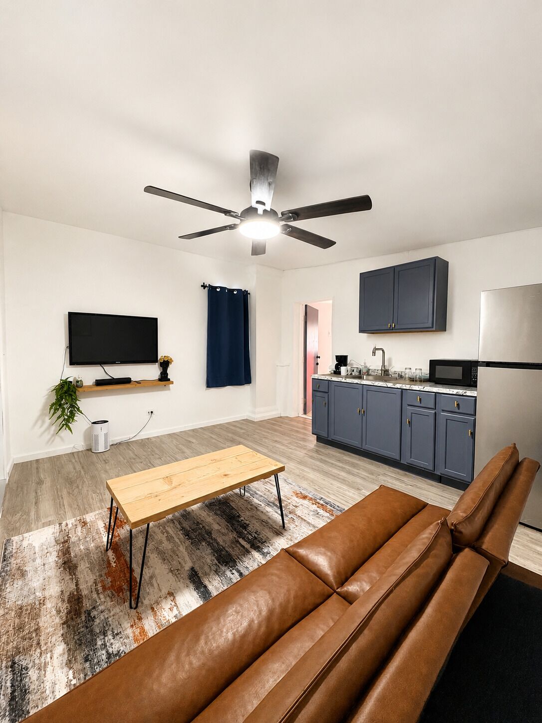 A modern living room with a sofa, a TV, a ceiling fan, and a kitchen area with cabinets and appliances.