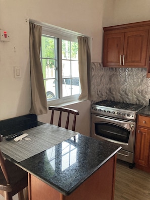 A kitchen with a granite countertop, wooden cabinets, a stainless steel stove, and a window with curtains. - Cozy studio in tranquil Ocho Rios with refreshing A/C with a wonderful loft bed! (Ocho Rios)