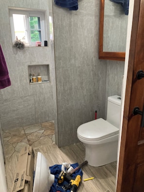 A bathroom with a toilet, a window, and a shelf with toiletries. - Cozy studio in tranquil Ocho Rios with refreshing A/C with a wonderful loft bed! (Ocho Rios)