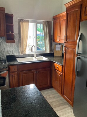 A kitchen with wooden cabinets, a granite countertop, a stainless steel refrigerator, and a window with curtains. - Cozy studio in tranquil Ocho Rios with refreshing A/C with a wonderful loft bed! (Ocho Rios)