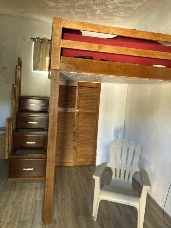 A bunk bed with a wooden frame, a red mattress, and a white plastic chair.