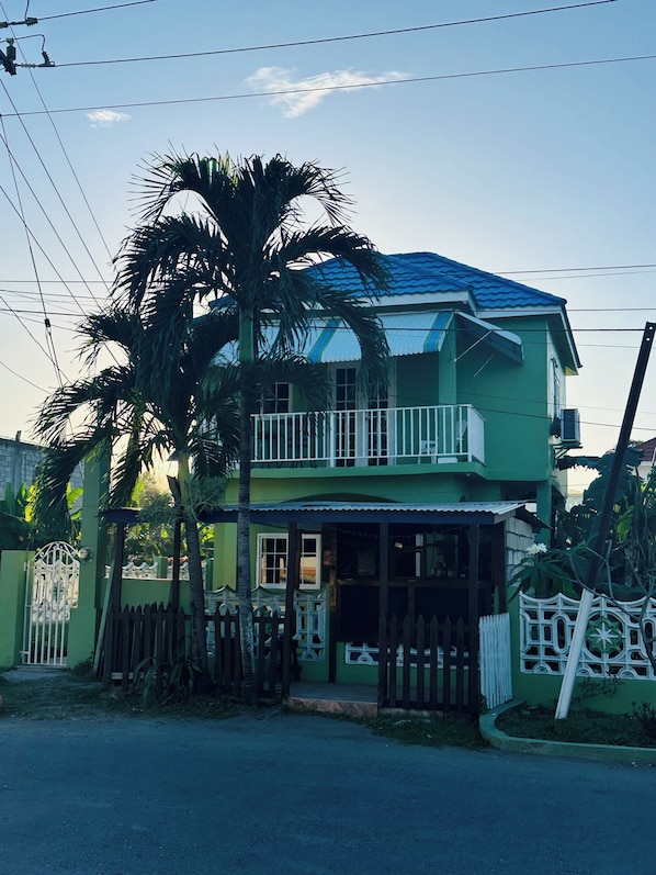 A two-story house with a balcony, a gated entrance, and a palm tree. - Cozy studio in tranquil Ocho Rios with refreshing A/C with a wonderful loft bed! (Ocho Rios)