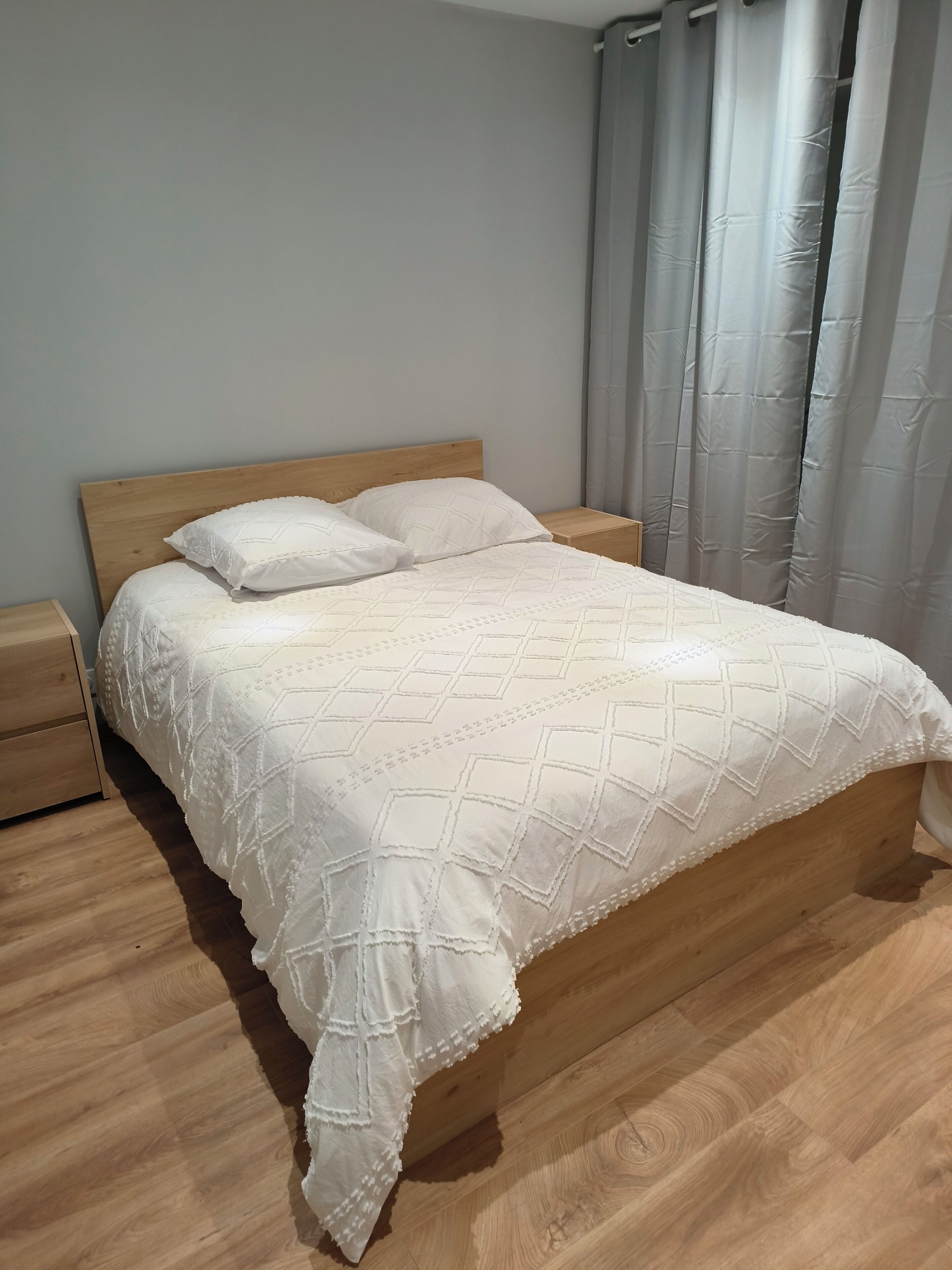 A neatly made bed with a wooden headboard, a wooden nightstand to the left, and a grey curtain to the right.. Charming renovated house