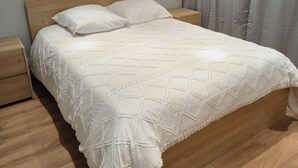 Una cama bien hecha con cabecera de madera, una mesita de noche de madera a la izquierda y una cortina gris a la derecha.