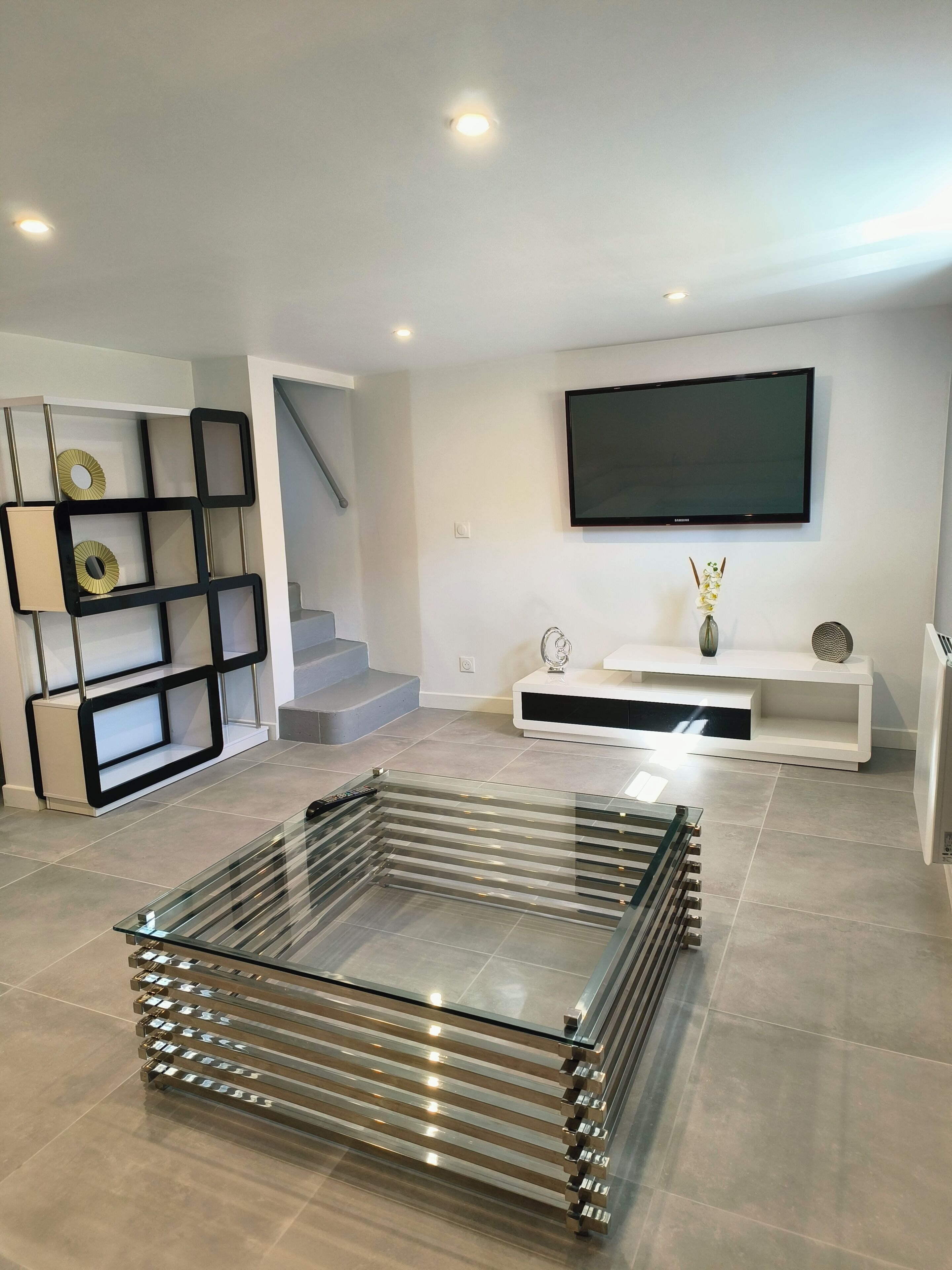 A modern living room with a glass coffee table, a flat-screen TV, and a minimalist shelving unit.