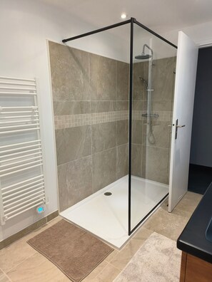 Een douchecel met glazen wanden, een bedieningspaneel, een witte radiator en een bruine mat.